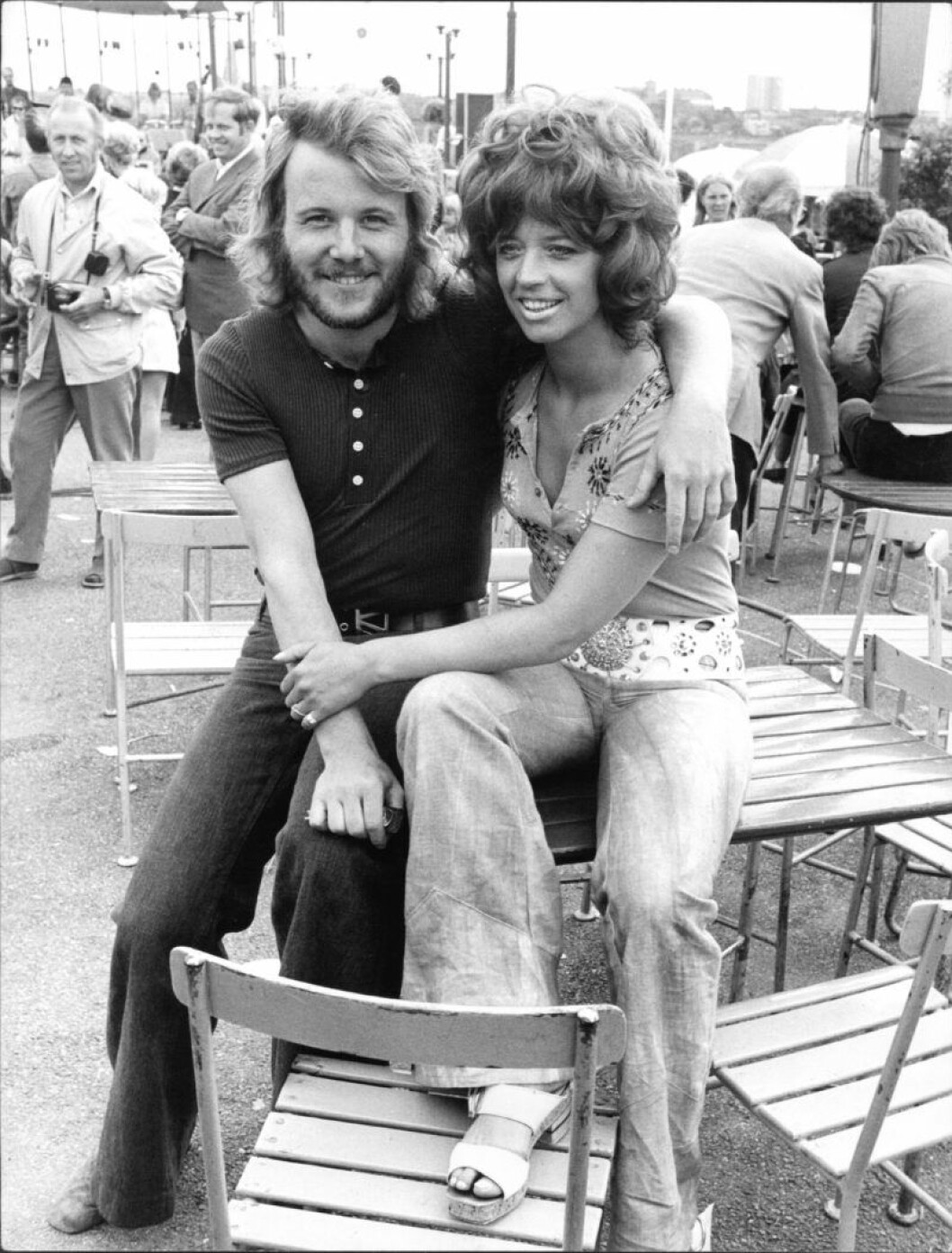 Benny Andersson och Lill-Babs sitter på ett bord och håller om varandra, med massor av folk i bakgrunden.