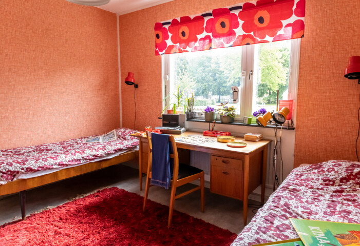 Här bor de fiktiva syskonen Lotta, 14, och Peter, 10. I fönstret hänger en gardin sydd i Marimekkotyget Unikko, som säljs än i dag. Sovrum i en lägenhet inredd med 70-talsdetaljer. Två sängar står mellan ett skrivbord. I fönstret hängen en gardin sydd i Marimekkotyget Unikko och på golvet en röd ryamatta.