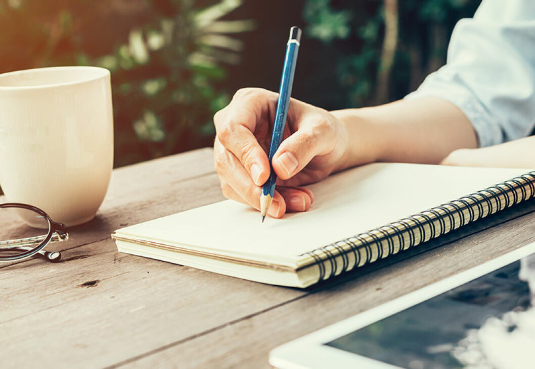 Bild på en hand som skriver med blyertspenna på ett skrivblock. Till vänster om blocket syns en vit kaffekopp och en bit av ett par glasögon. På höger sida syns en läsplatta.