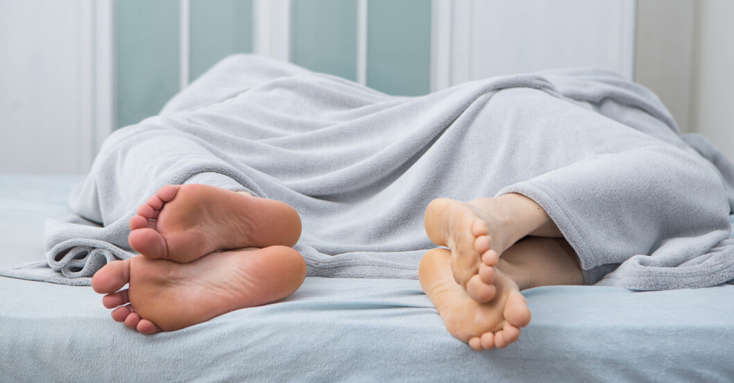 En man och en kvinna ligger under en grå filt på sängen, vända bort från varandra och det enda som syns är fötterna.
