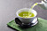 Grönt te hälls upp ur kanna ner i vit kopp.
