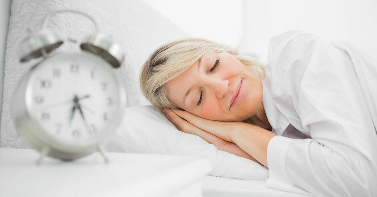 Väckarklocka i förgrunden och medelålders kvinna som sover i säng i bakgrunden