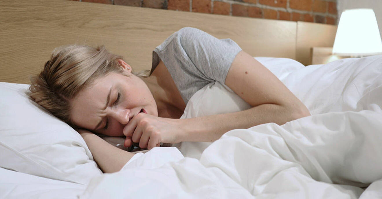 Blond kvinna med rynkad panna ligger i säng med vita sängkläder och håller knuten näve framför öppen mun.