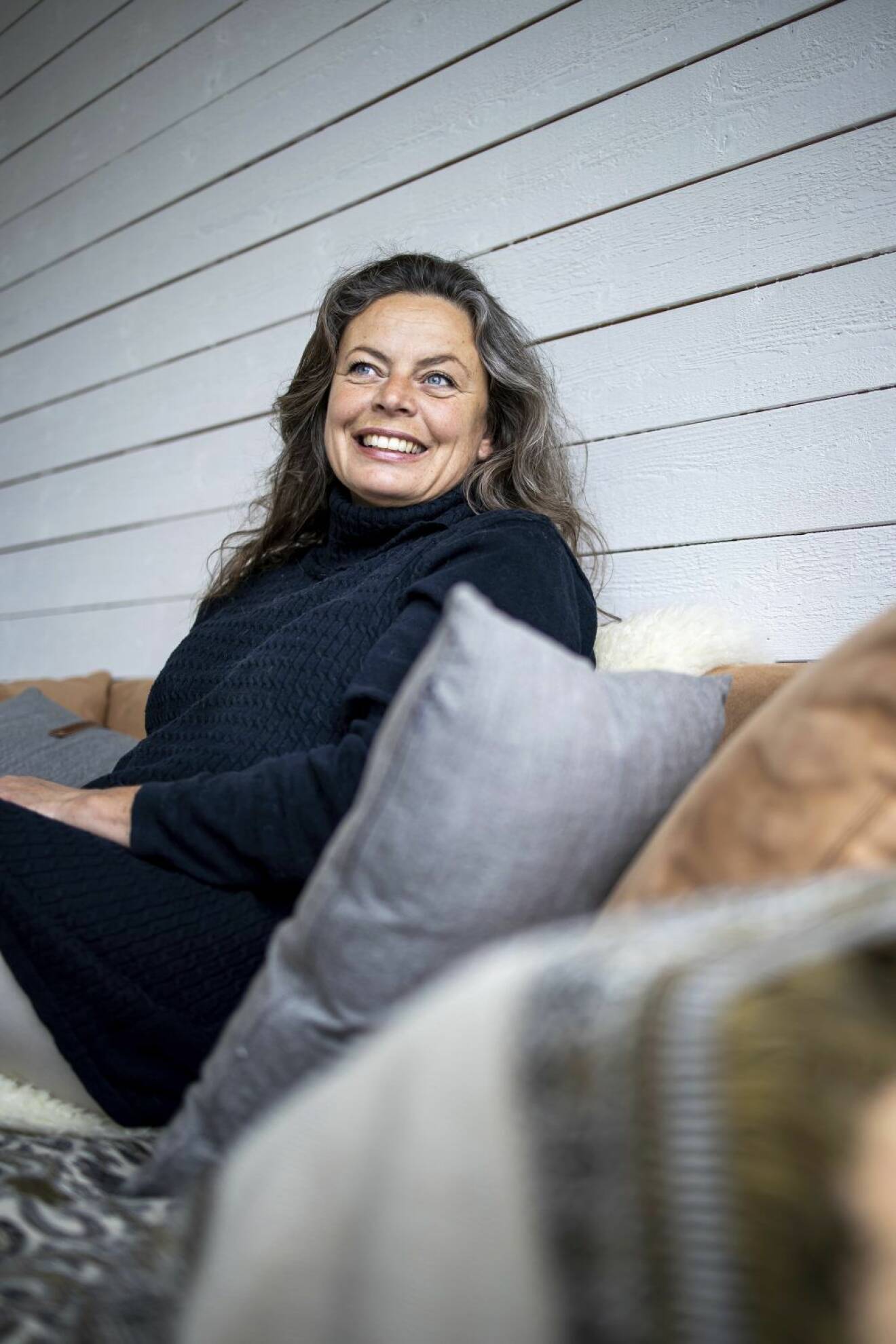 Kvinna med långt grått hår sitter i soffa mot kuddar mot trävägg och ler stort.
