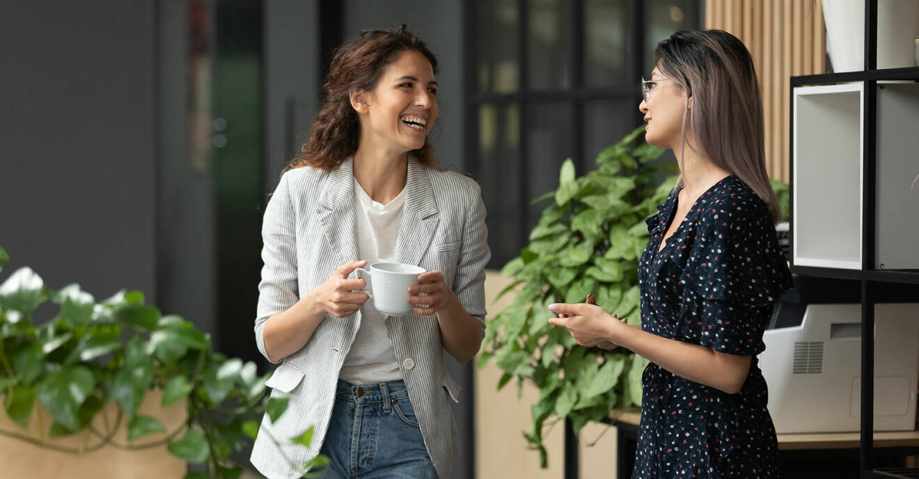 Två kvinnor står bredvid varandra, pratar och skrattar. Den enda har en kopp i handen.