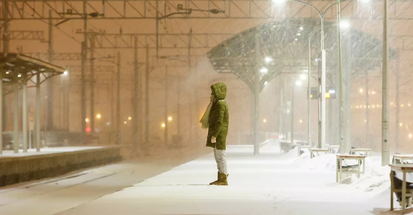Ensam kvinna i tjock jacka väntar på perrongen i mörkret och det snöar.