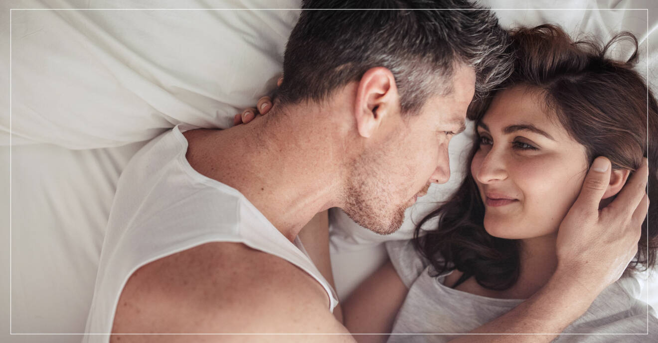 Man och kvinna ligger på säng med vita lakan och ser varandra i ögonen