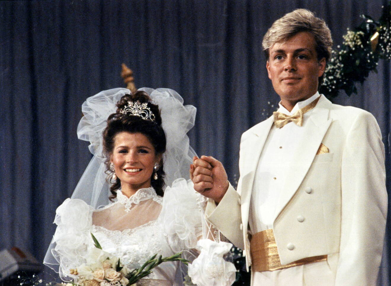 Carola Häggkvist och Runar Sögaard vid bröllopet 1990. Carola i en bröllopsklänning med tyll och spetsdetaljer och Runar i en vit smoking.