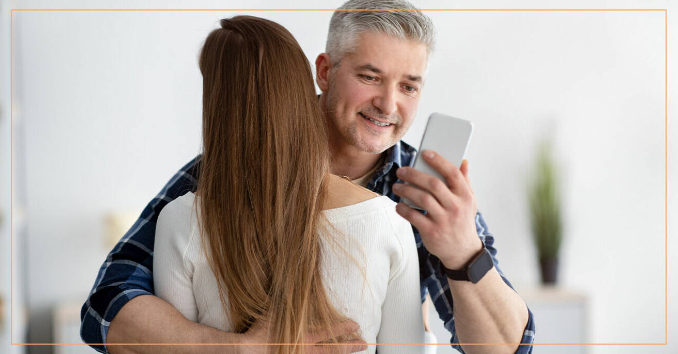 En medelålders man håller om sin partner medan han tittar i sin mobiltelefon bakom ryggen på henne