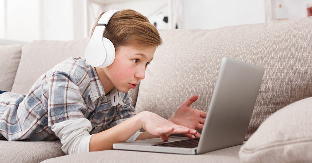Blond pojke med hörlurar ligger i soffa och skriver på dator