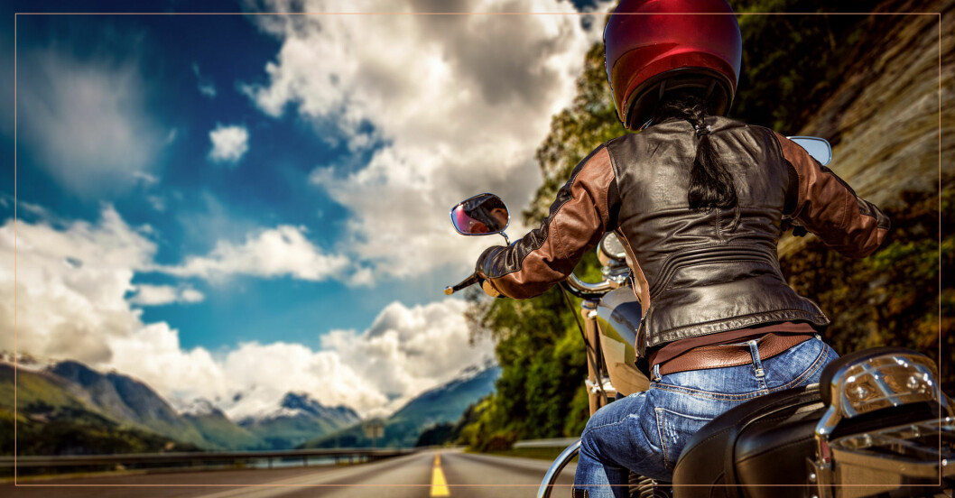 Kvinna i skinnjacka och hjälm kör motorcykel på väg genom vackert landskap.