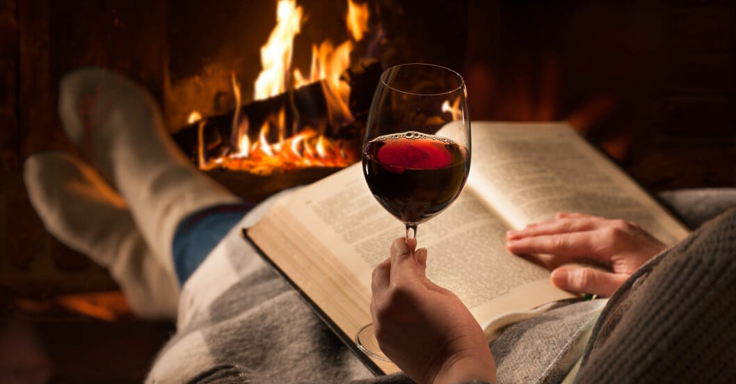 Kvinna läser en bok och dricker ett glas rödvin i lugn och ro framför brasan.