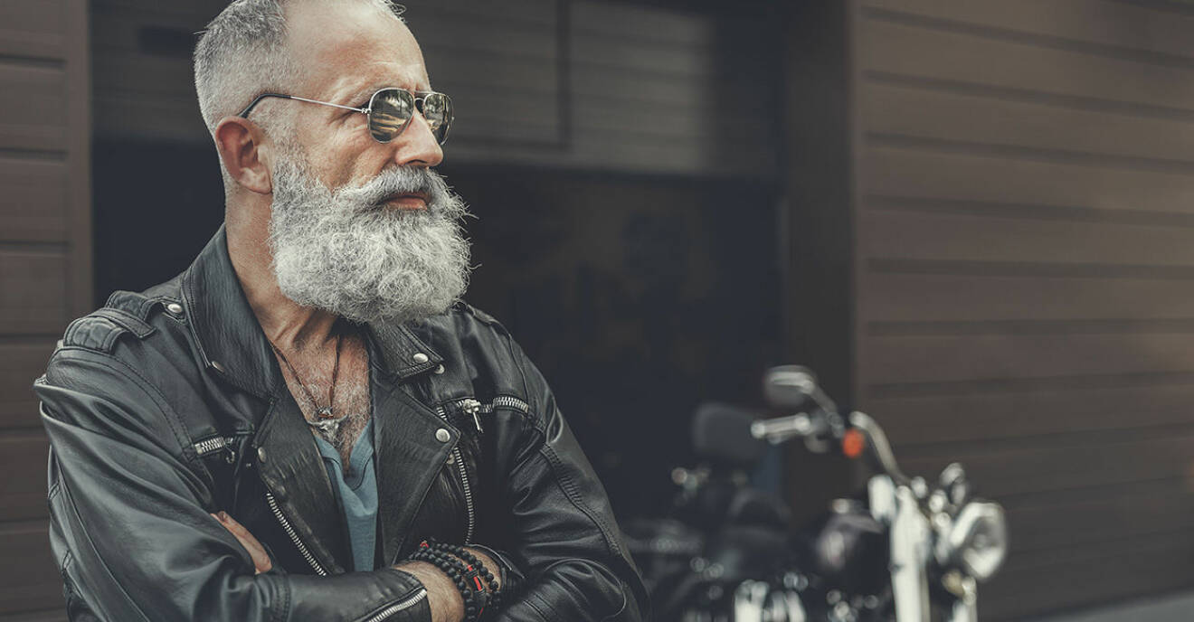 Äldre man med grått skägg och armarna i kors står framför en garageport och en motorcykel.